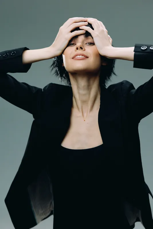 confident woman in black jacket with short hair posing with hands on head, stylish fashion portrait, studio background