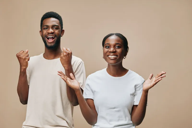 Joyful young Black couple expressing happiness and excitement with cheerful emotions, dressed in casual attire against a warm beige background