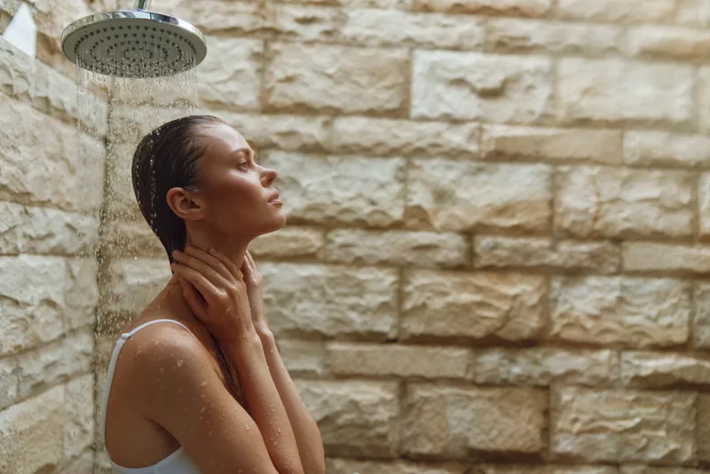 Woman relaxing under outdoor shower with stone wall background, water droplets, wet hair, and serene expression, wearing white swimsuit in natural light.
