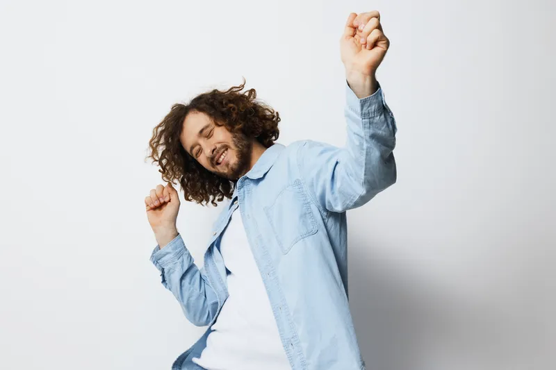 Happy young man with curly hair dancing joyfully in a light blue shirt against a white background, expressing positive emotions and carefree spirit