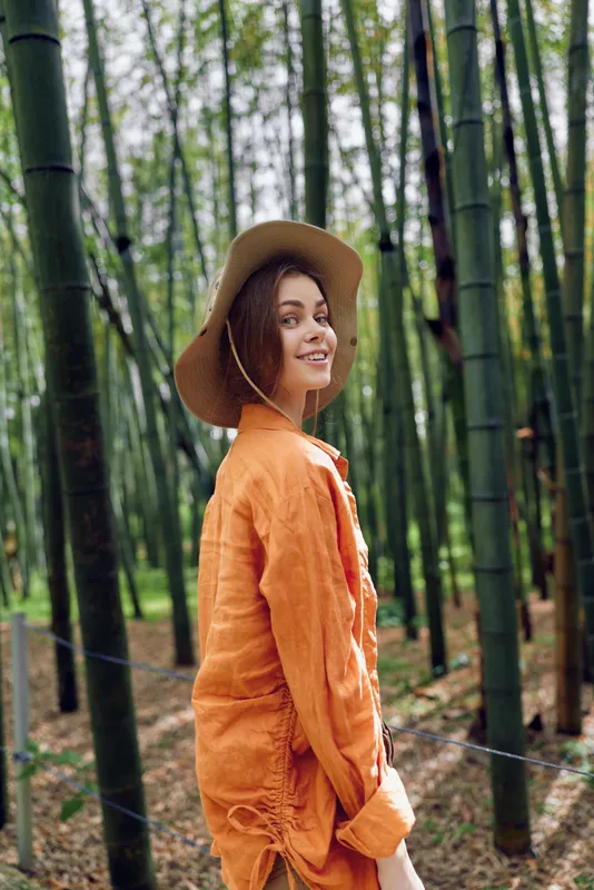 Woman in orange jacket and wide hat smiling in bamboo forest, portrait of a happy traveler surrounded by tall green bamboo and nature, casual outfit and relaxed outdoor walk.