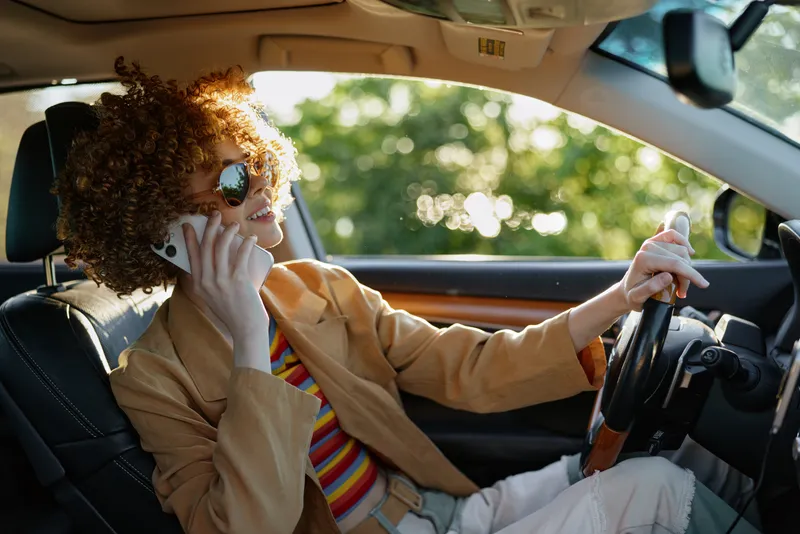 Woman in retro fashion sits in a car, talking on a phone, wearing sunglasses and a blazer, daylight streaming through windows, casual chic lifestyle moment on the road