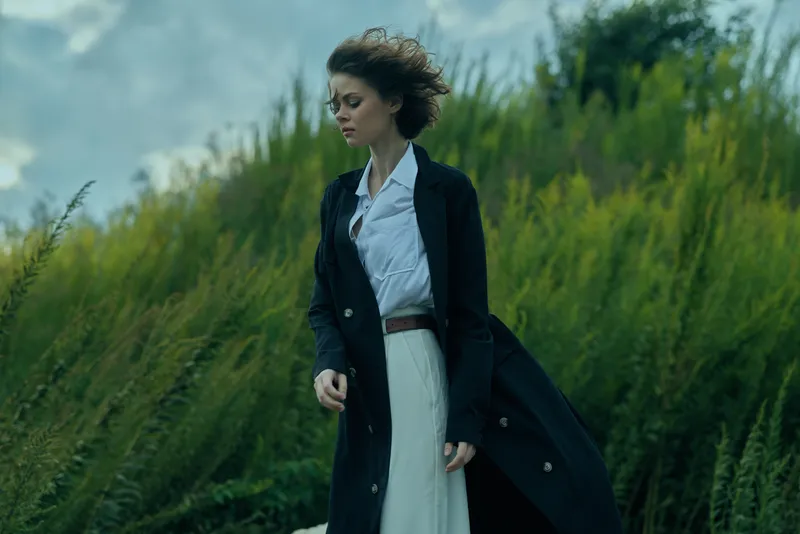 Woman in a dark coat over a light dress walks through a windy outdoor field, presenting professional style and determined expression amid nature, capturing motion and focus.