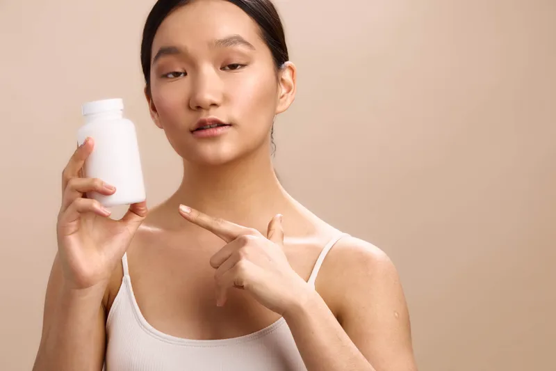 Asian woman holds a white bottle in a clean studio setting, presenting the product toward the camera with a calm smile, confident posture, and natural lighting, conveying health, beauty, and trust.