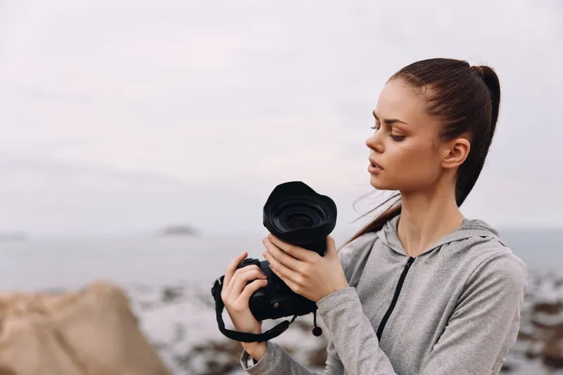 Young woman photographer in a gray hoodie holding a camera by the beach, showcasing focus and creativity, ideal for lifestyle and photography themes