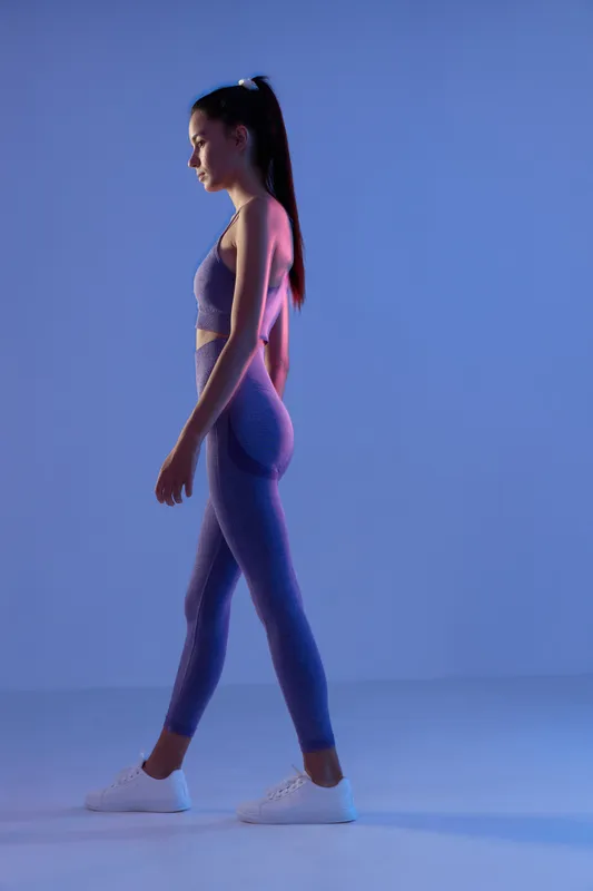 Sport fitness woman in purple activewear walking indoors with focused expression, tied hair, and white sneakers against a blue purple gradient background.