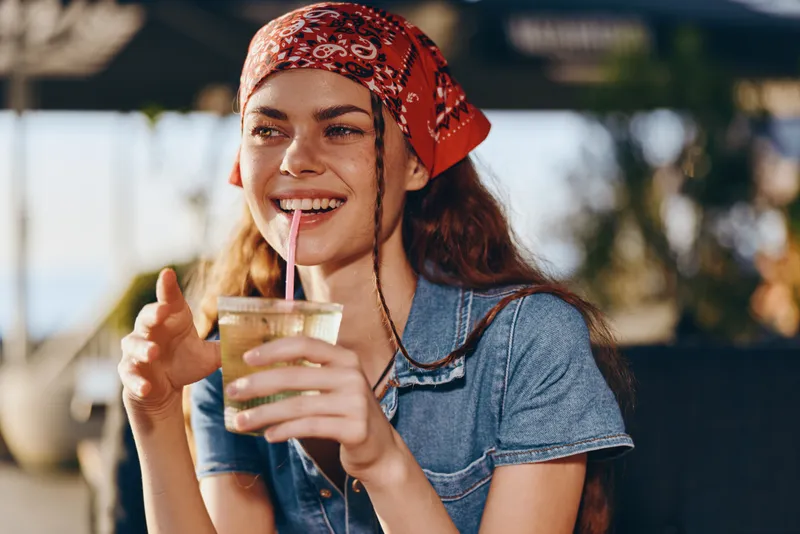 Smiling woman wearing a red bandana and denim shirt enjoying a refreshing drink outdoors with warm sunlight and relaxed summer atmosphere around her.