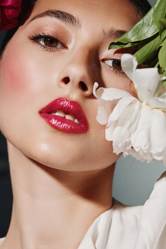 Close-up of young woman with glossy red lips and natural makeup posing with white flower against neutral background in creative fashion and beauty concept photo.