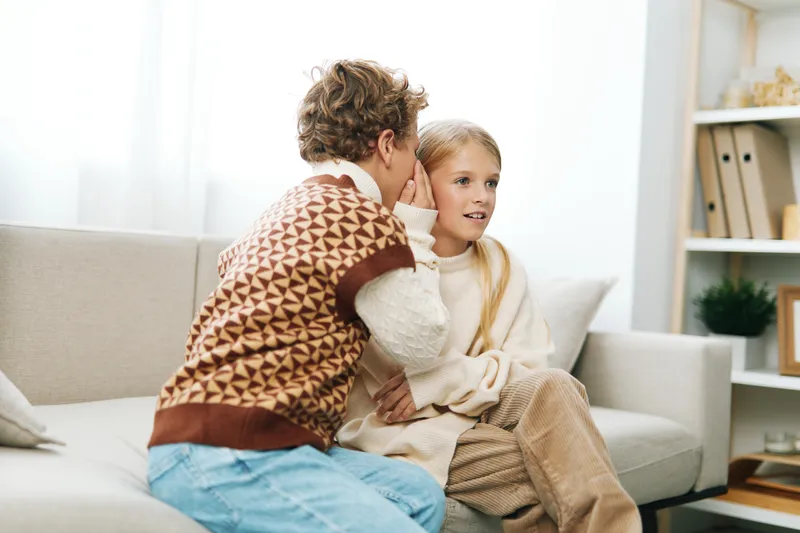Two children share a secret moment on a cozy sofa, leaning close as the boy whispers to the girl, a warm home scene filled with light and gentle mood.