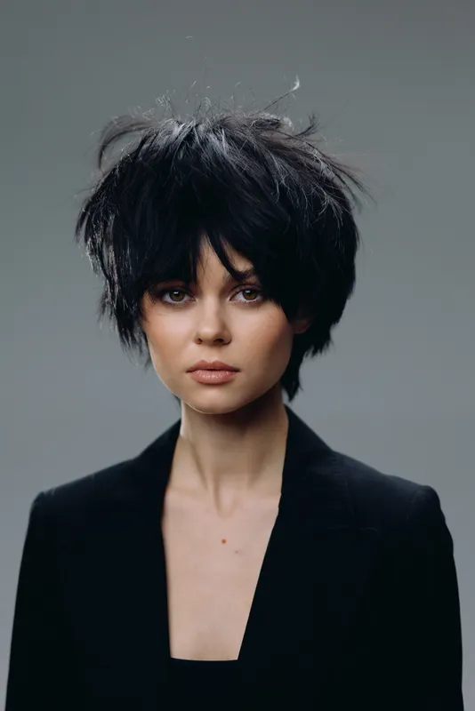 Portrait of a young woman with tousled black hair wearing a black blazer against a gray background