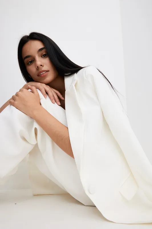 Young woman in white suit poses thoughtfully against minimalist white background, showcasing modern fashion with natural makeup and long dark hair.