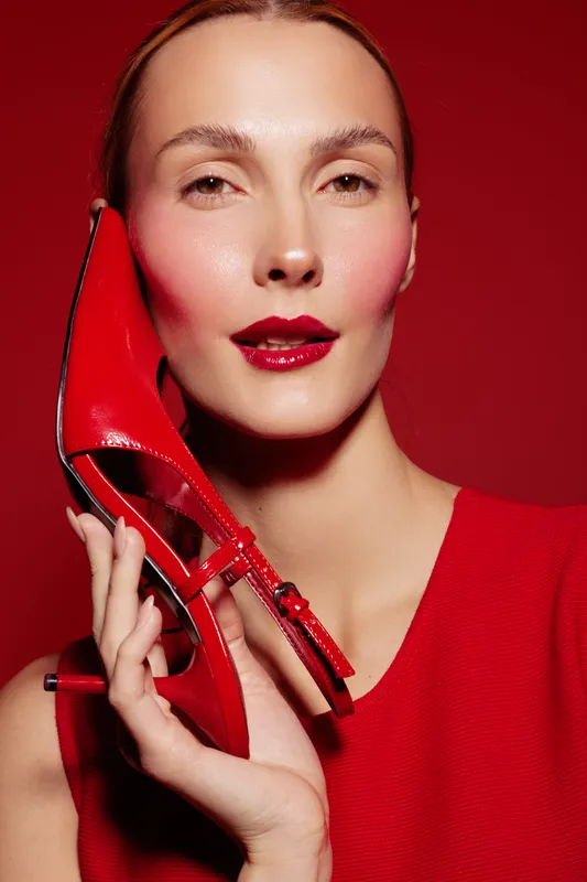 Close-up of a woman with glossy red lips holding a red high heel shoe near her face against a red background. Concept of fashion, beauty, and style with vibrant color contrast.