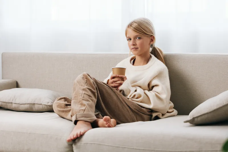 A young girl sits on a light sofa, holding a mug, relaxed and thoughtful, bathed in soft natural light. She wears a cream sweater and cozy pants, creating a warm home moment.