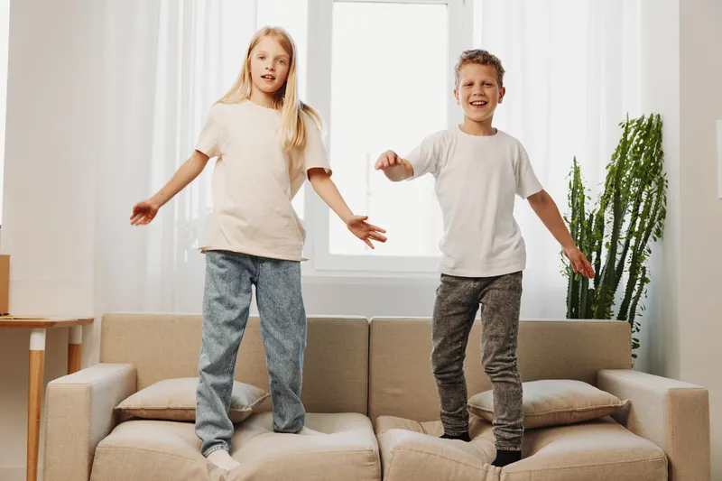 Two children standing on a sofa in a bright living room, smiling and playing together, casual clothes and home environment, concept of childhood joy and family fun.