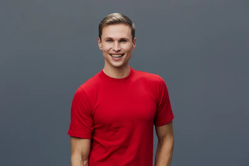 Smiling young man with short brown hair wearing a bright red t-shirt looking at camera with a cheerful expression against plain gray background. People lifestyle concept