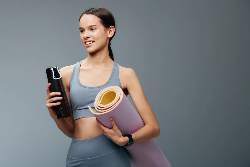 Athletic woman in a sports bra and leggings holds a water bottle and a rolled yoga mat, ready for a workout. Clean studio background highlights fitness and sport focus.