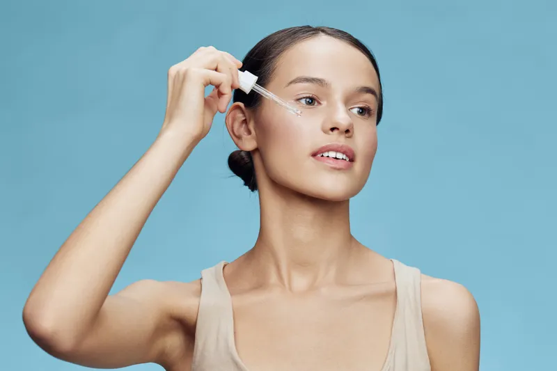 Young woman applying serum to her skin, featuring natural beauty and glowing complexion, against a light blue background, ideal for skincare promotions