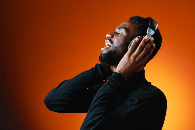 Young Black man enjoying music with headphones, expressing joy and creativity against a vibrant orange background, perfect for lifestyle and music themes