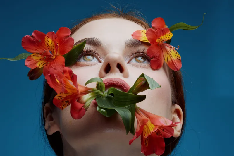 Young model with red flowers on her face, showcasing beauty and nature against a blue background, evoking emotions of serenity and freshness