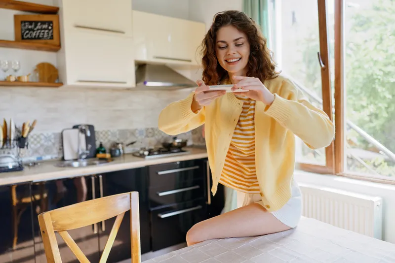 Relaxed home lifestyle scene featuring a smiling woman in a bright kitchen, sitting on a counter, using a smartphone, casual clothes, warm natural light, everyday moment