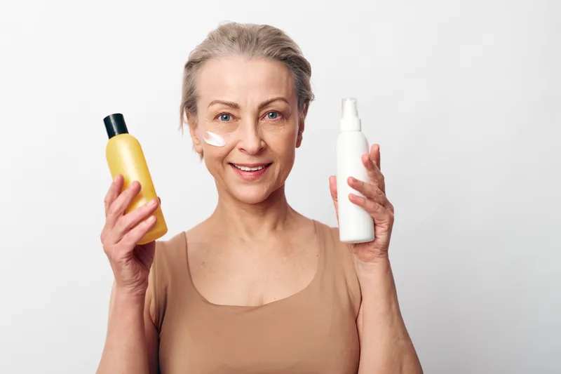Smiling mature woman with natural skin holds two cosmetic bottles, applying cream on face. Concept for skincare, beauty routine, anti aging, and self care at home.