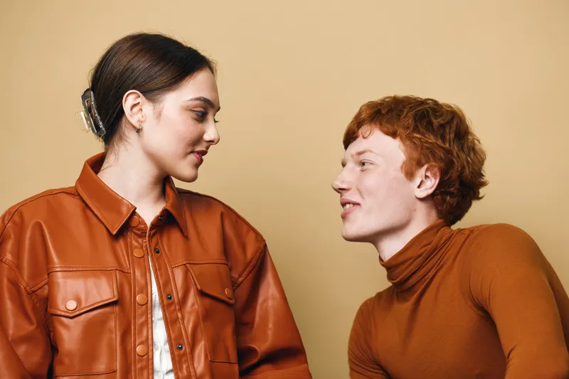 Young man and woman looking at each other with smiles in casual brown outfits. Indoor portrait with warm beige background showing friendship and connection between people.