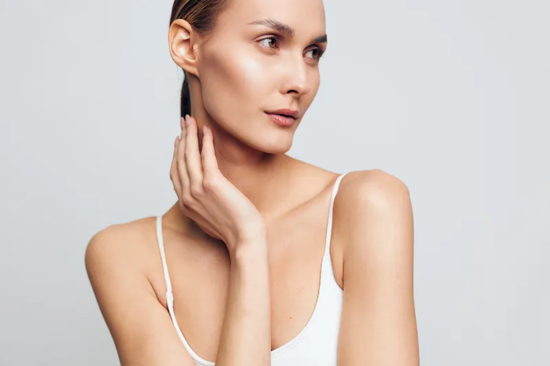 A young woman with clear skin and natural makeup gently touches her neck while looking to the side. She wears a white tank top against a plain light background. Concept for beauty and skincare.