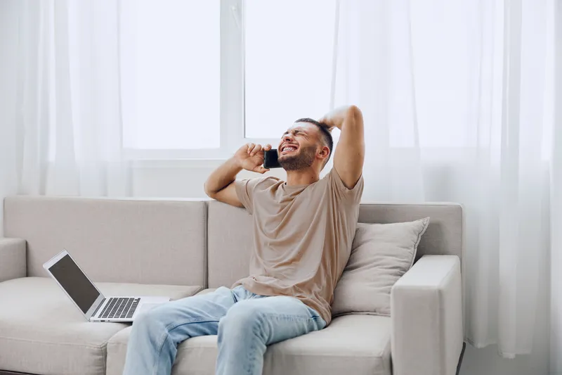 Happy man talking on the phone with laptop nearby, relaxed atmosphere, bright light coming through curtains, modern living room setting, joyful moment captured.