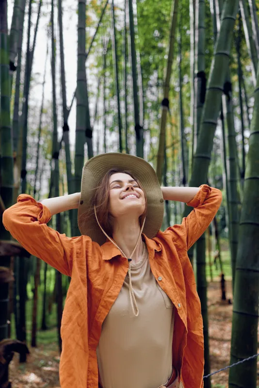 Woman in straw hat and orange jacket standing in bamboo forest, smiling with eyes closed and arms raised, enjoying nature, relaxation and outdoor travel in a peaceful green grove.