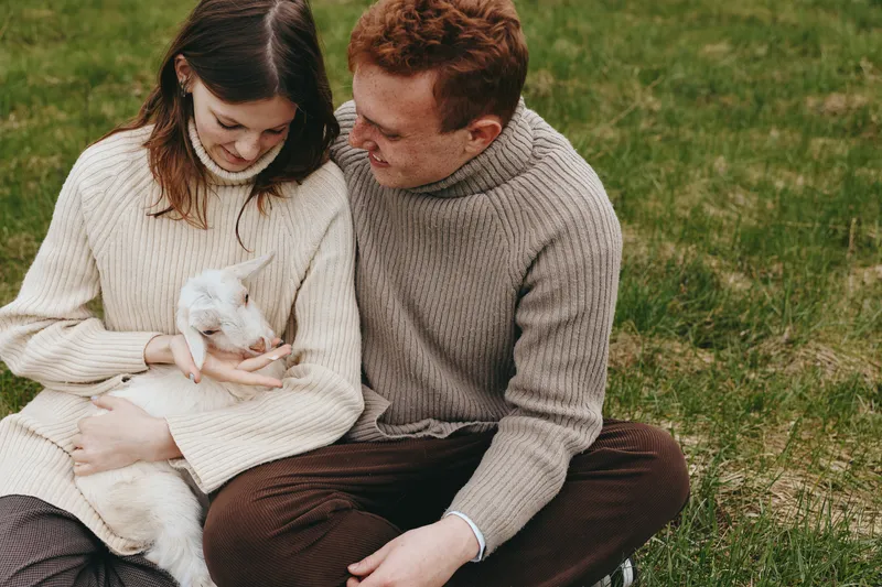 Couple in cozy knitwear bond outdoors with a goat kid, capturing a warm lifestyle moment of affection and family. Soft daylight on grass creates a gentle, tender mood with companionship.