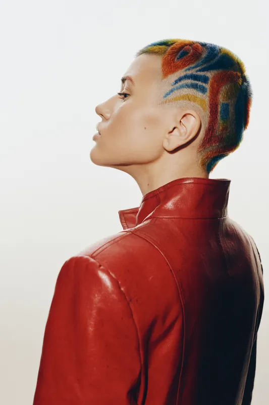 Profile of a woman with colorful shaved hair wearing a red leather jacket, showcasing bold hairstyle and fashion in a studio setting.
