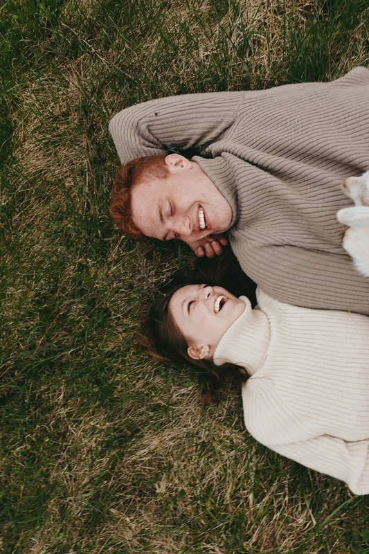 Smiling couple lying on grass in warm sweaters, enjoying a carefree outdoor moment, sunlight and soft shadows around them, lifestyle photography capturing comfort, connection, and wellbeing.