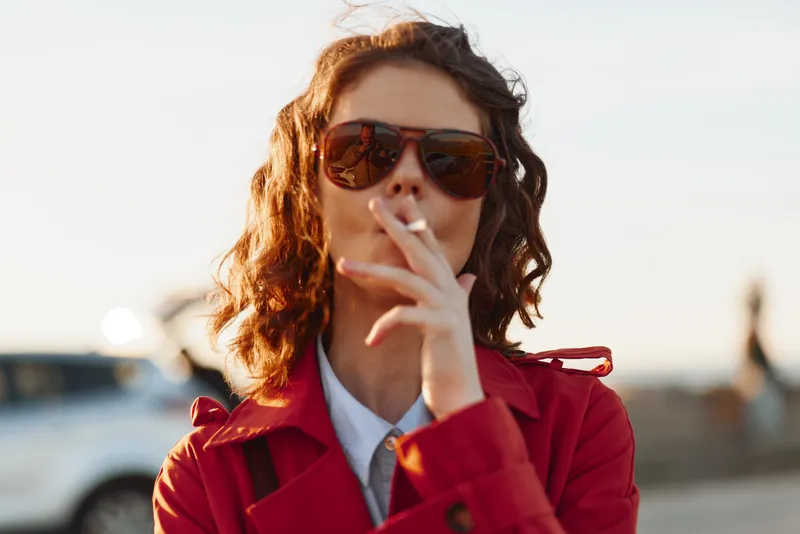 Woman wearing sunglasses and a red coat smokes outdoors in a sunlit portrait, conveying a stylish urban vibe and confident, casual mood.