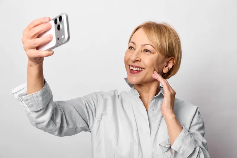 Smiling elderly woman selfie with smartphone, portrait in studio, touching her cheek and face, communicating with friends, clean neutral background, joyful modern mood.