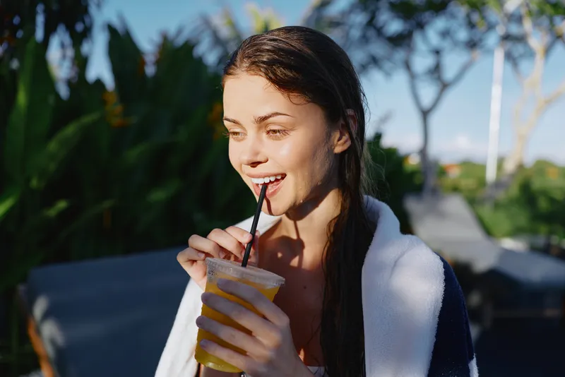 A woman outdoors with a towel over her shoulders smiles while sipping a cold beverage through a straw, capturing a lifestyle moment of refreshment, relaxation, and wellness in bright daylight.