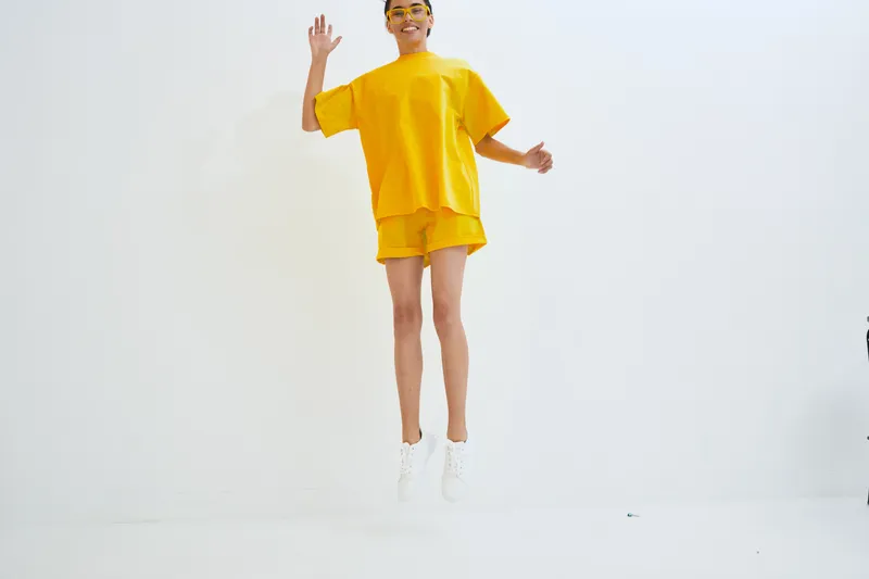 Young woman jumping happily in bright yellow casual outfit with white sneakers against clean white studio background, expressing joy and energy.