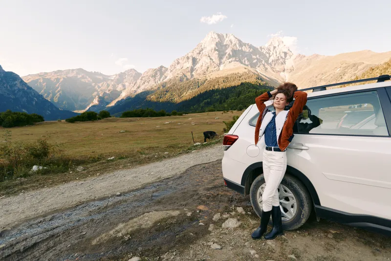 Woman by car near mountains and a gravel road, travel portrait in scenic landscape and nature. Young female leaning on SUV enjoying relaxation, freedom and outdoor adventure.