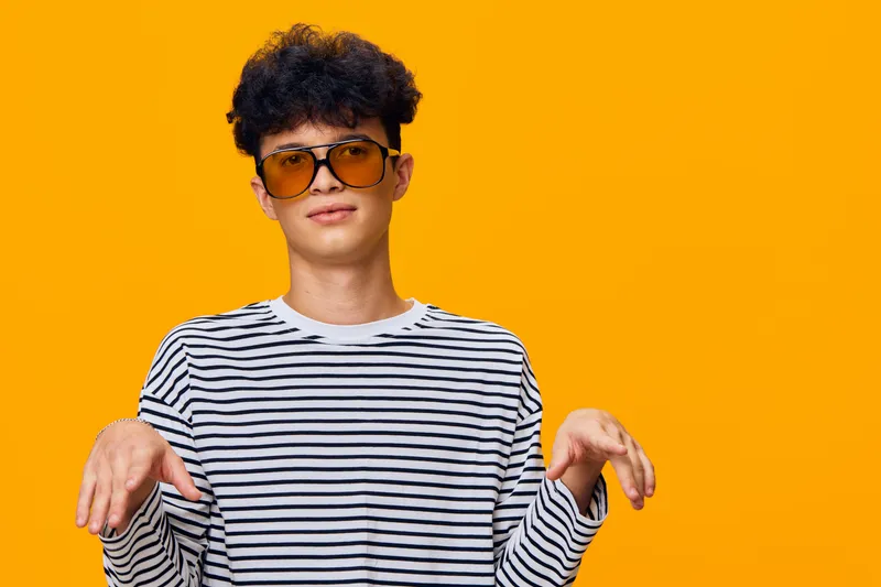 Young man with curly hair wearing large sunglasses and striped shirt poses playfully against a vibrant orange background, showcasing casual style and fun attitude.