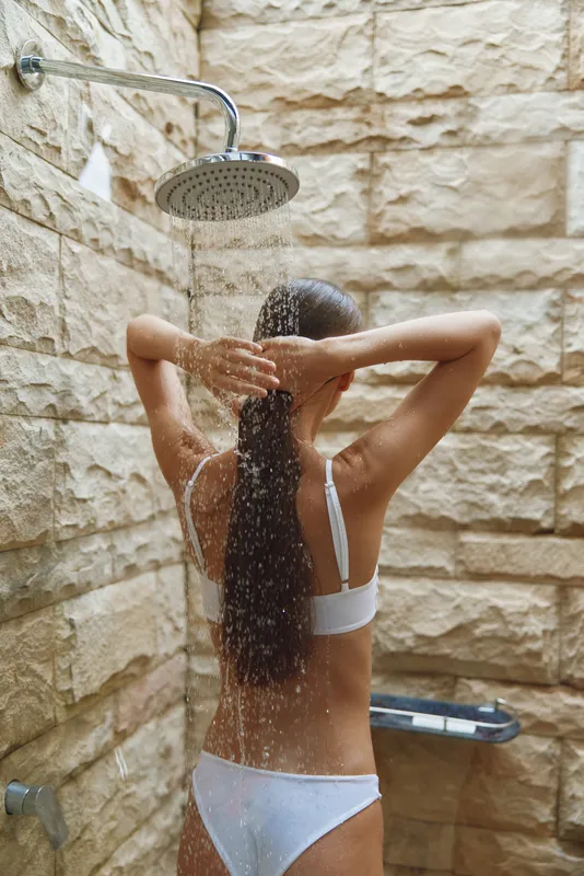 woman showering outdoors under rain showerhead with natural stone walls, wet long hair, white bikini, relaxing moment, summer freshness
