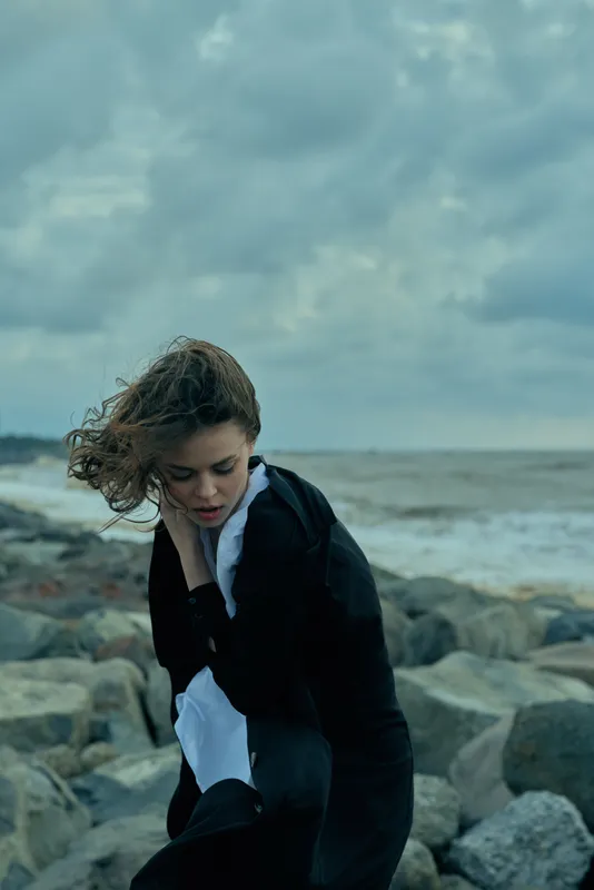 Woman in a black coat and white shirt on a windy coast, stepping over rocks by the sea, capturing a dramatic moody moment of movement and spray against a cloudy sky
