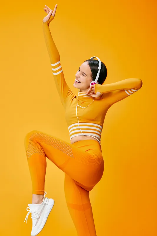 Energetic woman in orange athletic wear dancing with headphones, performing a high kick mid jump against a vibrant orange background, showcasing fitness, sport, movement and rhythm.