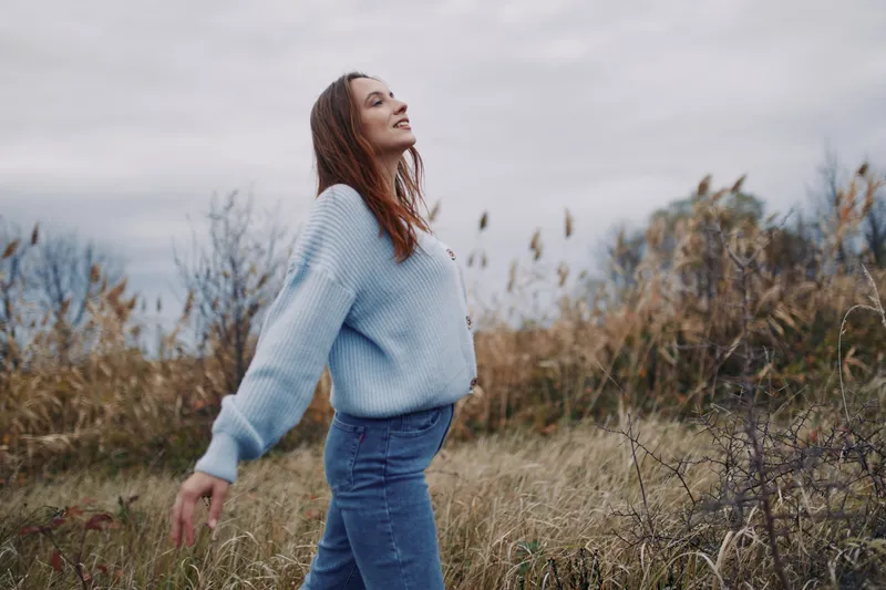 A candid portrait of a woman in a soft blue knit sweater walking through a natural field, conveying authenticity and credibility through relaxed posture and genuine mood.