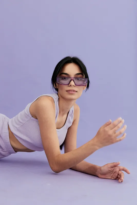 Young woman wearing purple sunglasses and lilac tank top lies on lavender background, showcasing modern fashion and relaxed vibe with stylish accessories.