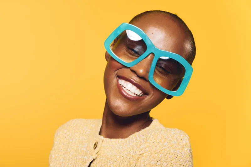 Smiling African woman with dark skin wearing large blue sunglasses and a yellow sweater poses against a bright yellow background. Concept of fashion, joy, and vibrant style.