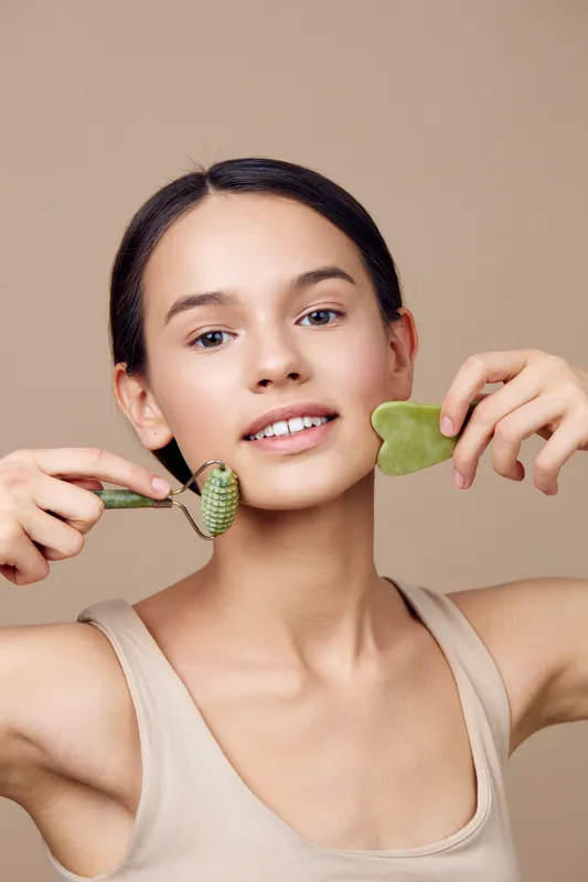 Young woman practicing self care using jade roller and gua sha tool for facial massage, promoting healthy skin and relaxation with a natural look