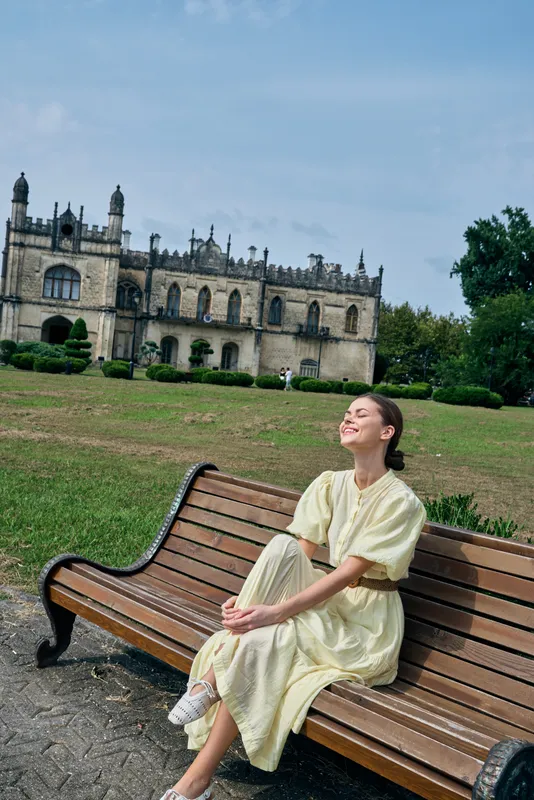 woman sits on a bench in a park, smiling softly, as a grand historic castle with ornate architecture rises in the background, creating a calm travel scene today for photography