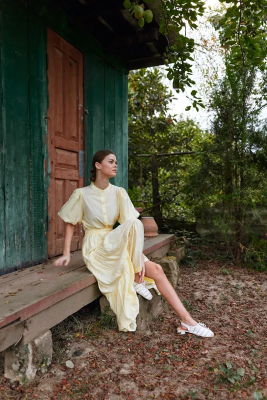 woman dress porch cottage nature relaxation, young model in yellow vintage outfit seated on rustic wooden bench by green cabin, authenticity inclusivity