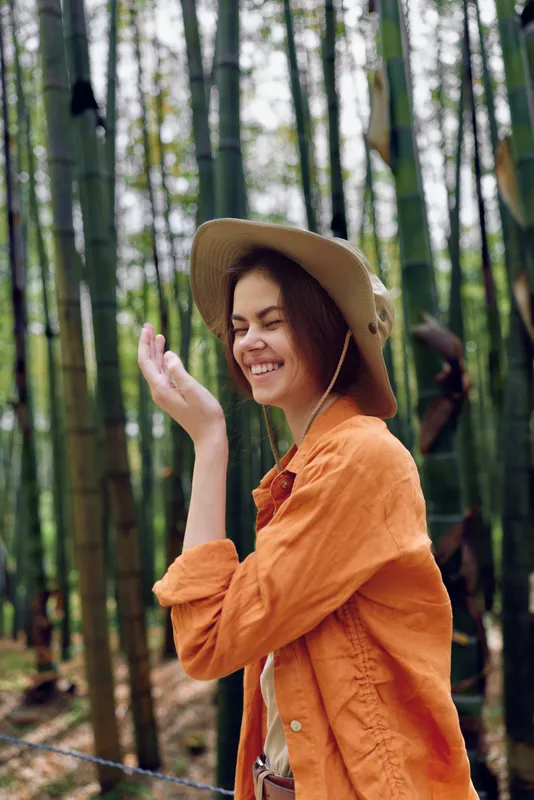 Woman smile in bamboo forest wearing a wide hat and orange jacket, portrait of happy traveler enjoying nature and outdoor adventure with candid laughter in a serene green grove.