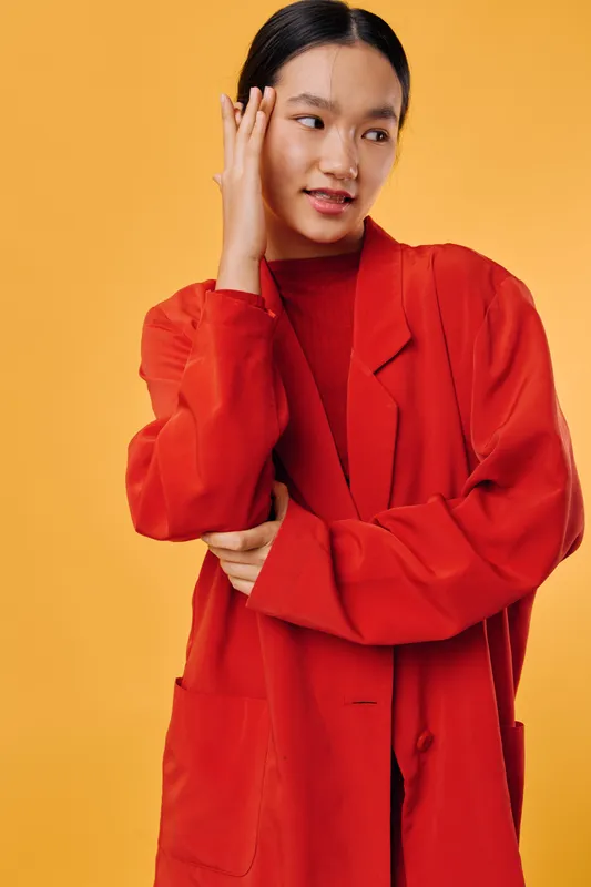Young woman wearing red coat and turtleneck posing with hand near face against orange background. Fashion, beauty, casual style with confident expression and smooth hair.