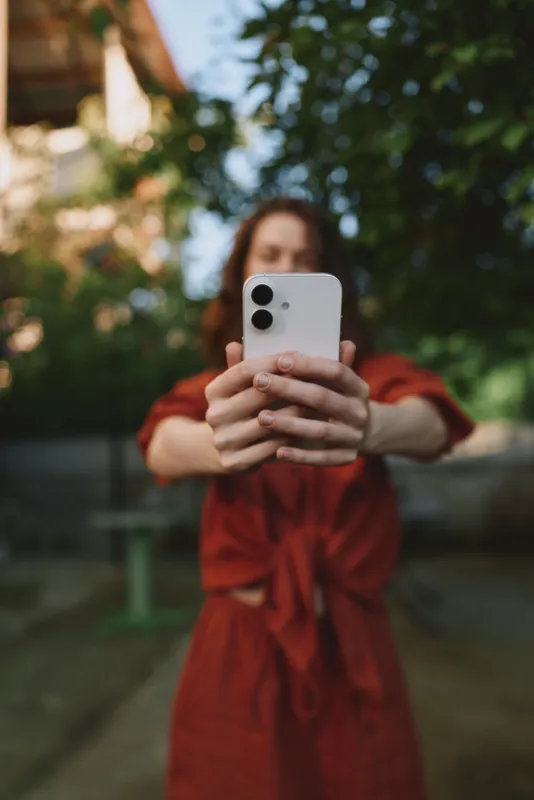 Woman lifestyle portrait outdoors, a woman in a red dress takes a selfie with a smartphone in a sunlit park, trees in background, casual pose, candid and relaxed mood